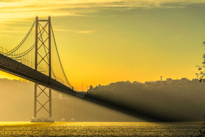 Low angle view of 25 de abril bridge over river against sky during sunset