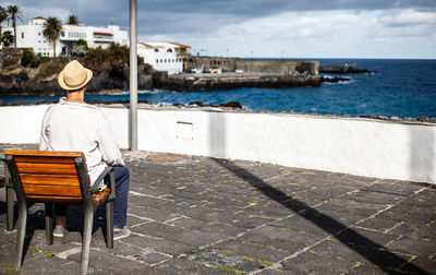 Rear view of man sitting on bench at observation point by sea against sky