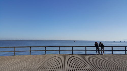 People standing on railing by sea against clear sky