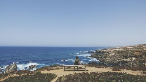 Rear view of boy sitting on bench against clear sky