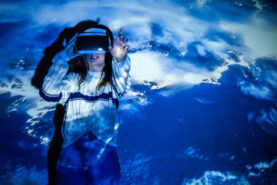 Young woman wearing virtual reality simulator standing against blue background