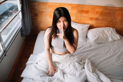 Portrait of young woman with finger on lips sitting on bed