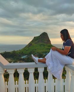 Woman sitting on railing against sky during sunset