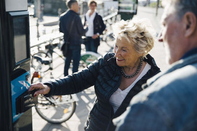 Senior woman operating bike vending machine by man on sidewalk in city