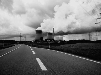 Country road by silos emitting pollution against cloudy sky