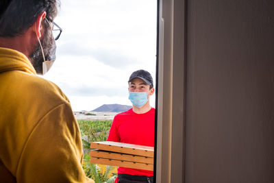 Smiling delivery man in mask holding pizza while standing at door