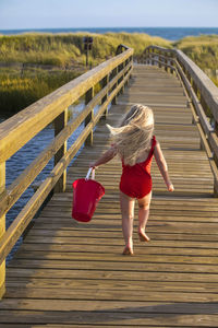 Little girl from behind running on bridge to beach with red bucket