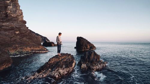 Full length of woman standing on rock by sea against clear sky during sunset