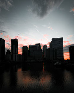 Silhouette buildings by river against sky during sunset