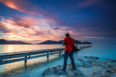 Rear view of man standing at beach against sky during sunset