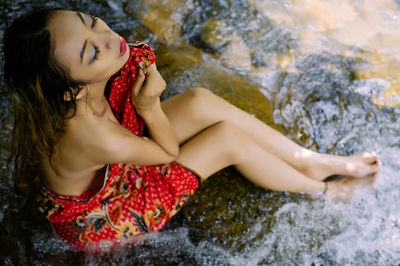 Sensuous woman wrapped in red fabric sitting on rock formation by flowing stream