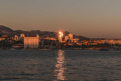 Illuminated buildings by sea against sky during sunset
