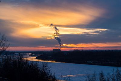 Silhouette of factory by sea against sky during sunset