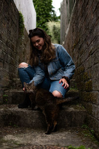 Full length of young woman against wall stroking a cute cat. 