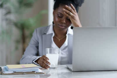 Ill woman holding glass with medicine by laptop on table