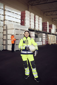 Portrait of female blue-collar worker in protective workwear holding hardhat while standing at industry