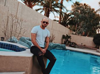 Portrait of young man wearing sunglasses while sitting by swimming pool