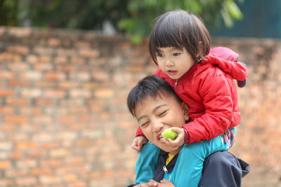 Happy smiling vietnamese boy eating an apple and holding his little sister sitting on neck