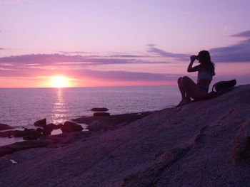 Side view of woman sitting on rock at beach against sky during sunset