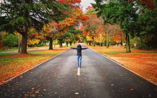 Rear view of woman standing with arms outstretched on road during autumn