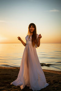 Beautiful young woman in a white long dress near the sea at sunset stands