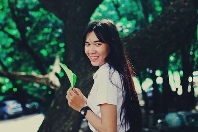 Portrait of smiling young woman holding leaf standing against trees