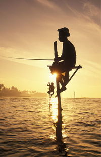 Low angle view of silhouette man stilt fishing in sea against orange sky