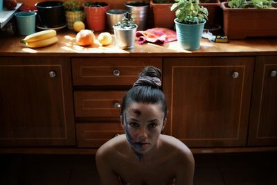 High angle portrait of shirtless young woman with messy make-up sitting in kitchen at home