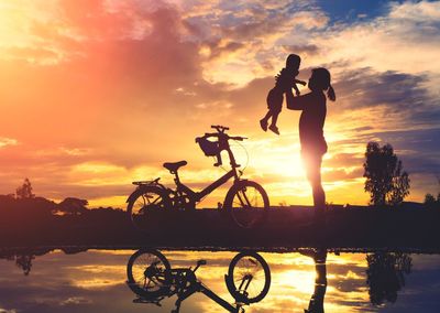 Silhouette of mother and daughter with bicycle against sky during sunset