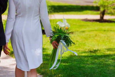 Rear view midsection of groom by bride holding bouquet on footpath