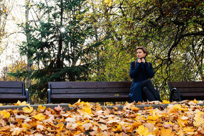 A woman wraps herself in a coat sitting on a bench in an autumn park