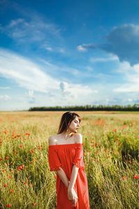 A girl in a red dress above the knee stands in a poppy field during the day against the sky