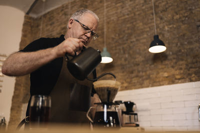 Low angle of concentrated mature male barista in apron adding water from kettle through paper filter while brewing pour over coffee at counter