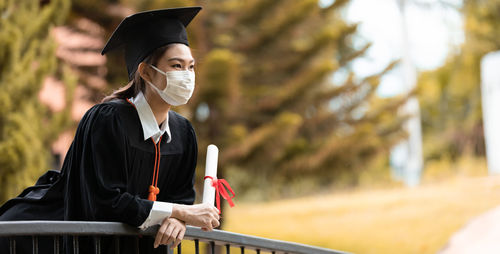 Woman in graduation gown holding certificate against trees