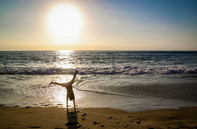Rear view of woman practicing cartwheel on shore at beach against sky during sunset
