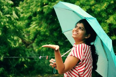 Side view of smiling young woman with umbrella while standing against tree during monsoon