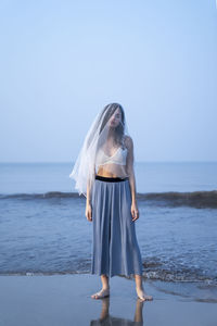 Woman wearing veil standing at beach against clear sky