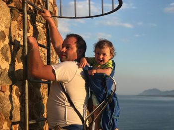 Father climbing ladder on wall while carrying son against sky