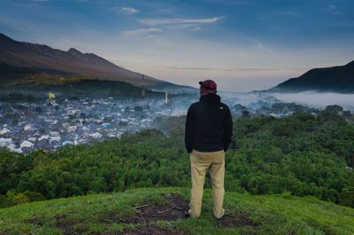 Rear view full length of man standing on mountain against cityscape during foggy weather