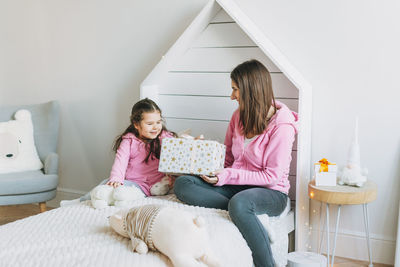 Young mother and cute girl in pink family looks hoodie on bed in children room, bright scandinavian 