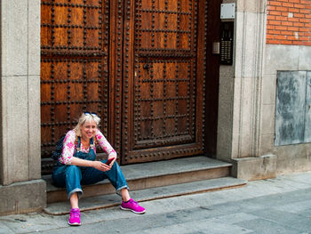 Full length of smiling woman holding mobile phone while sitting at entrance