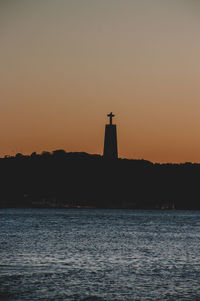 Silhouette lighthouse by sea against orange sky