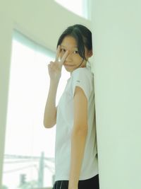 Portrait of woman showing victory sign while standing by white wall