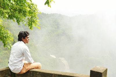 Rear view of man sitting on railing against mountain during foggy weather