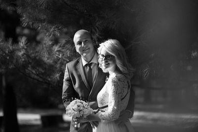 Portrait of bride and bridegroom standing against trees at park