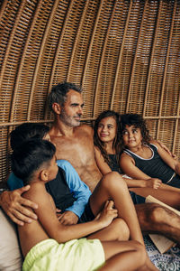 Shirtless father sitting with arm around children in wicker hut at beach