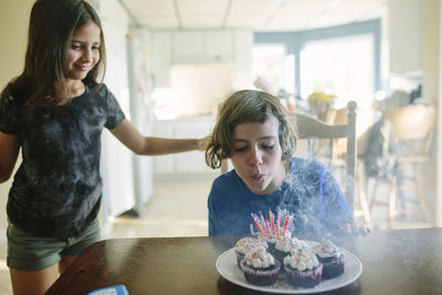 Smiling girl looking at sister blowing birthday candles on table at home