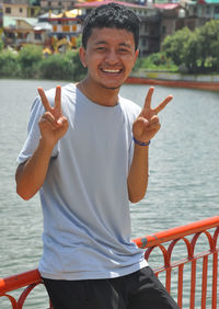 A aisan young guy smiling posing by lake with making victory hand sign and looking at camera