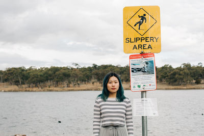 Portrait of young woman standing by warning sign at lakeshore against sky