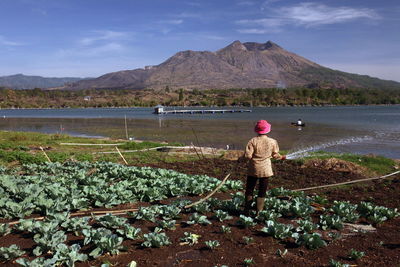 Rear view of farmer working at riverbank by mountain against sky